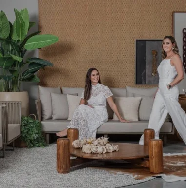 Duas mulheres vestidas de branco, uma em pé com a mão no bolso e outra sentada em um sofa
